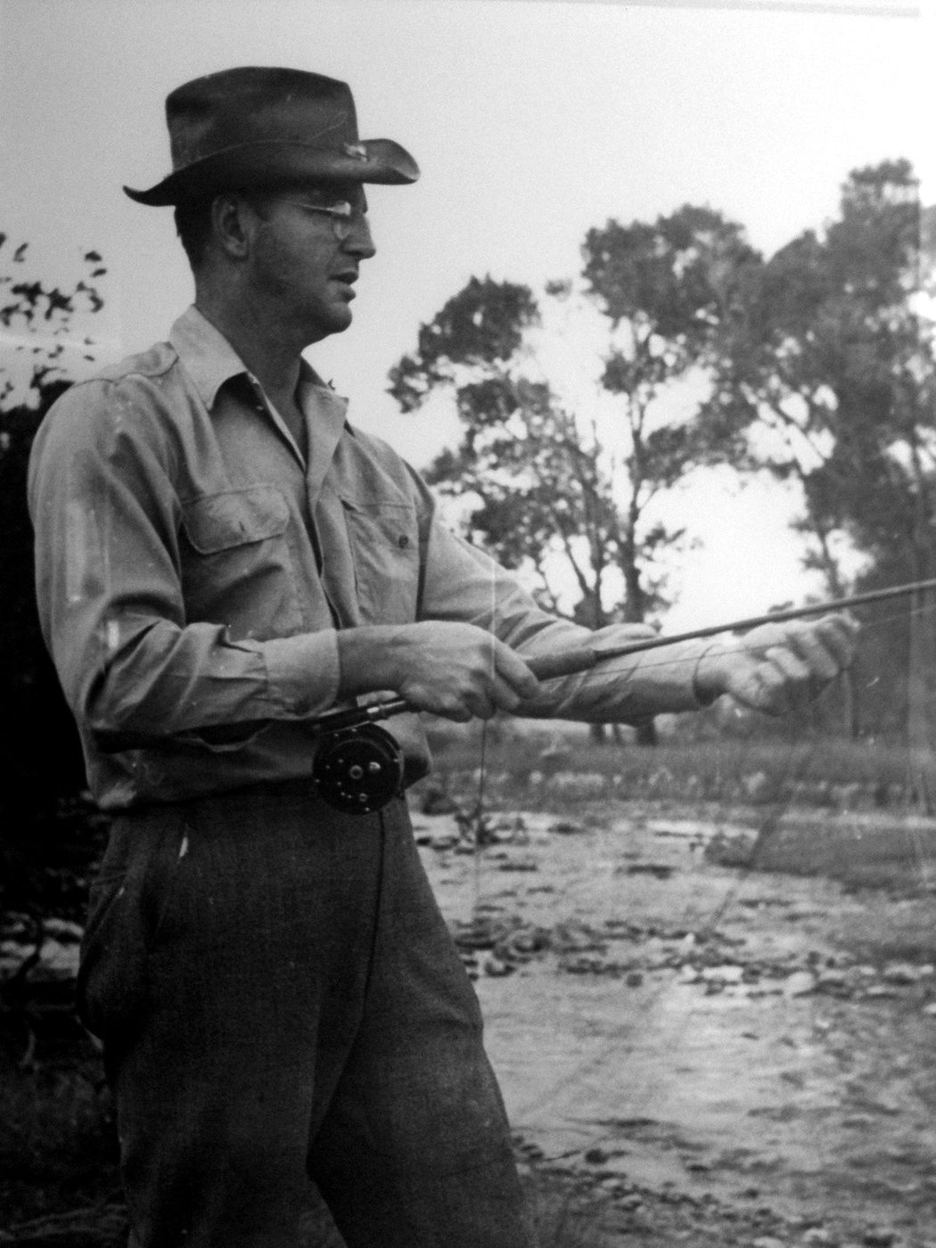 Frank Seney in a hat, holding a fly rod beside a stream
