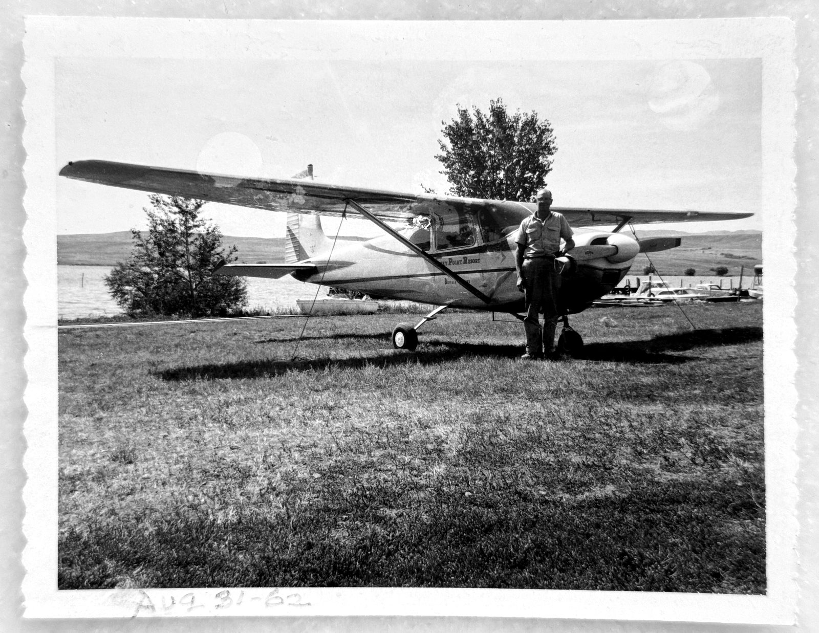 A high-wing airplane lettered Seney Point Resort on a grass strip, with a man standing beside it